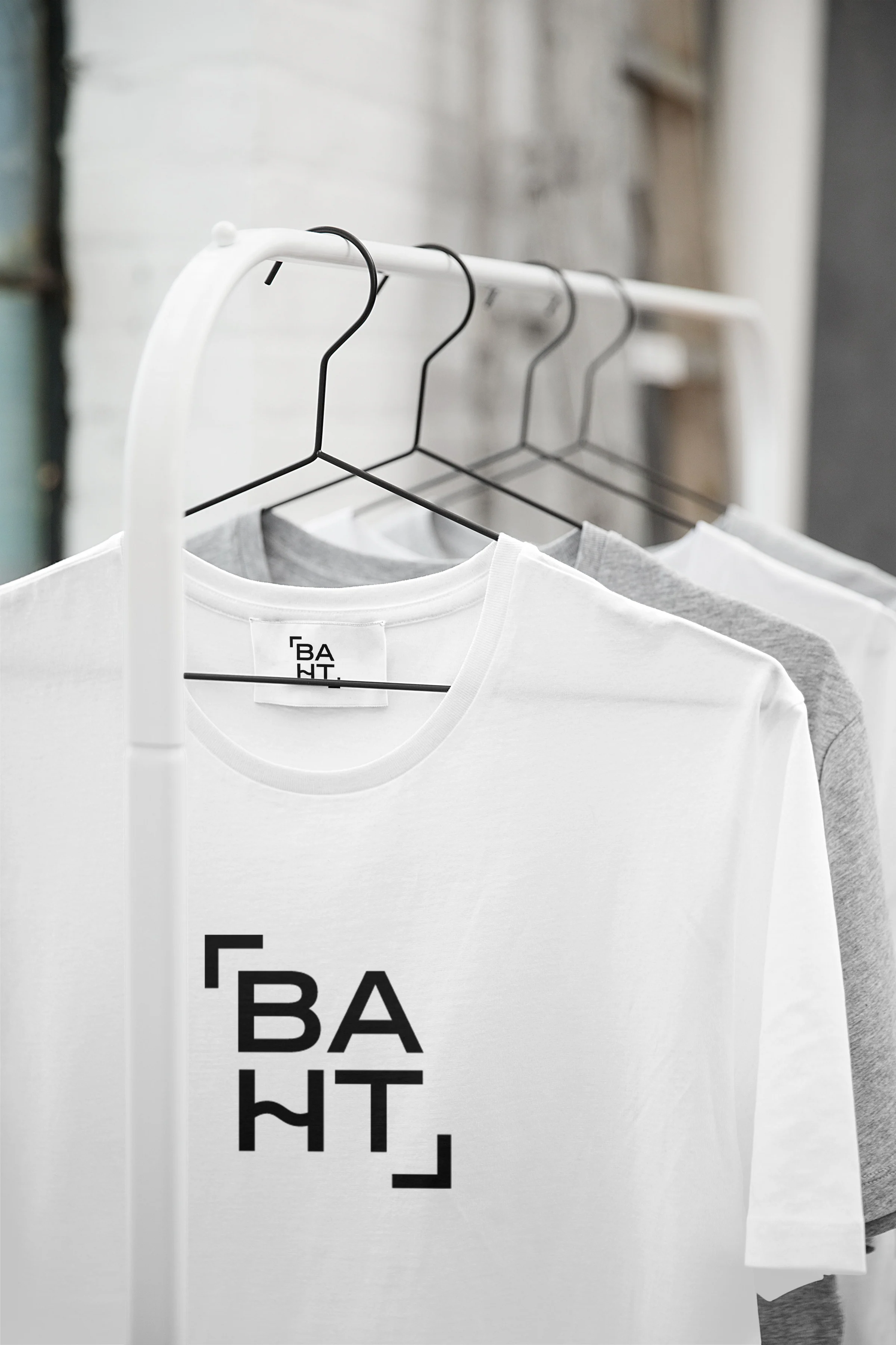 White and grey T-shirts on a rail with black hangers, the front one printed with a large baht logo and a logo neck label.