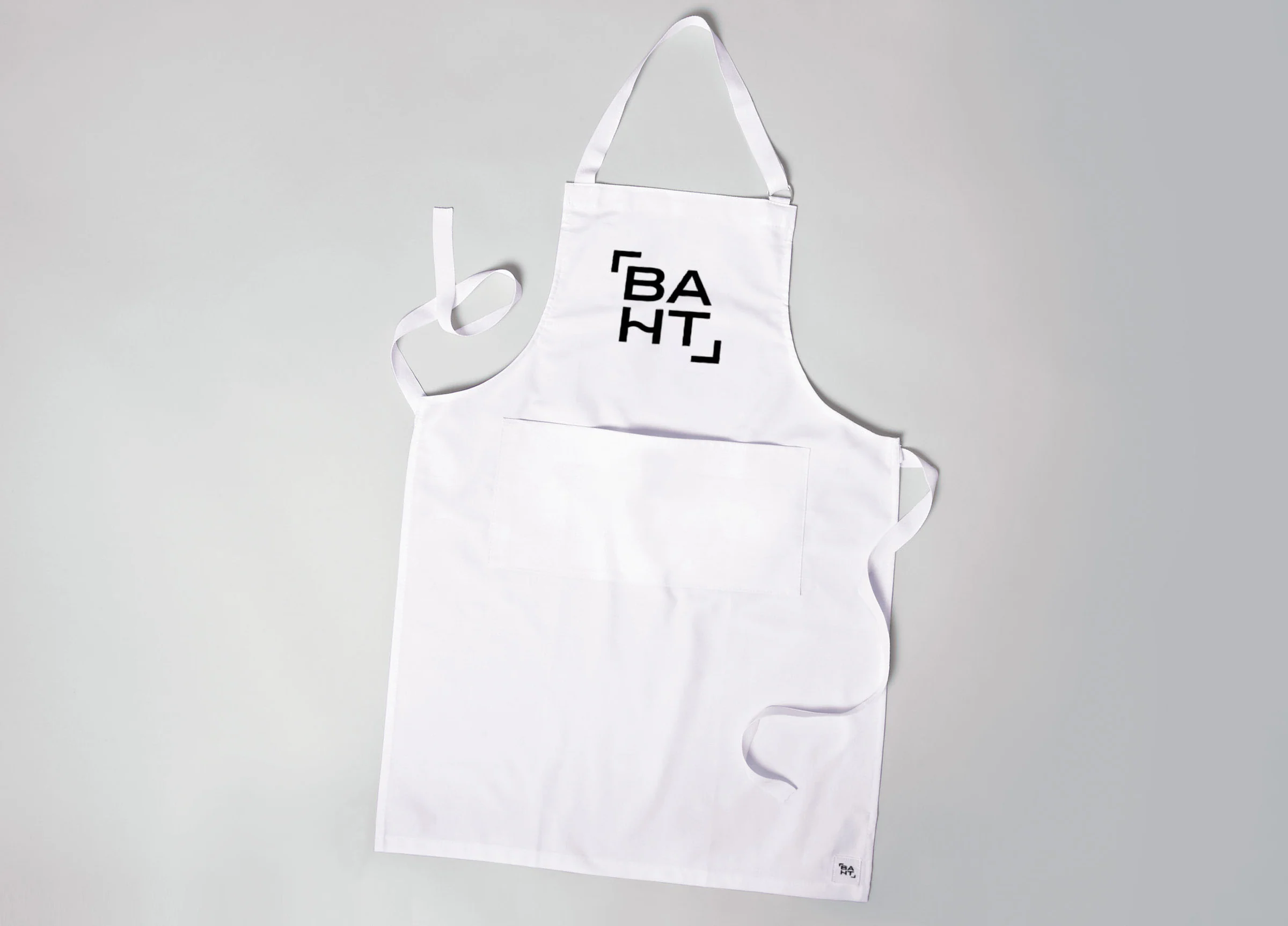 White apron with the baht logo on the bib and a small logo tag at the hem.