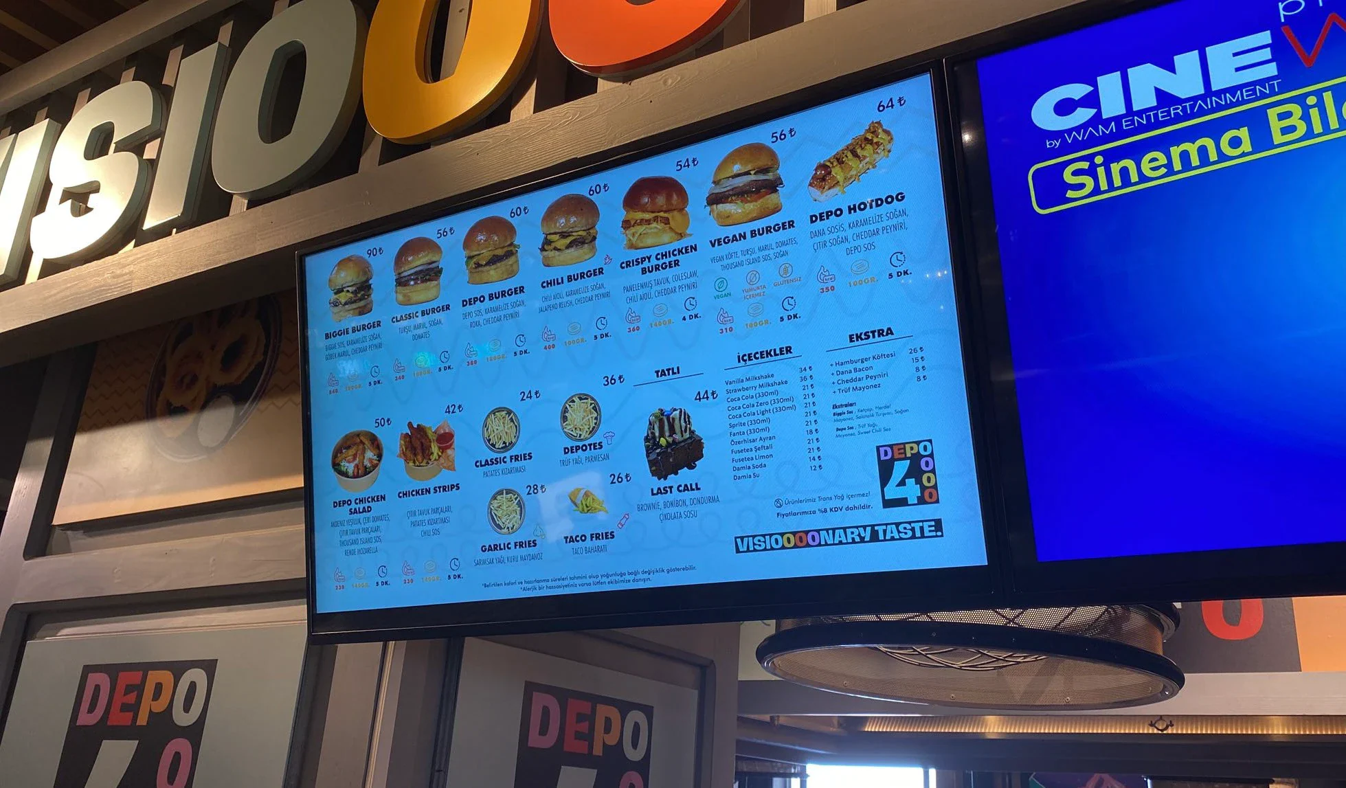 Light blue digital menu board with burger photos and prices, mounted under the lettered sign beside part of a CineWAM screen.