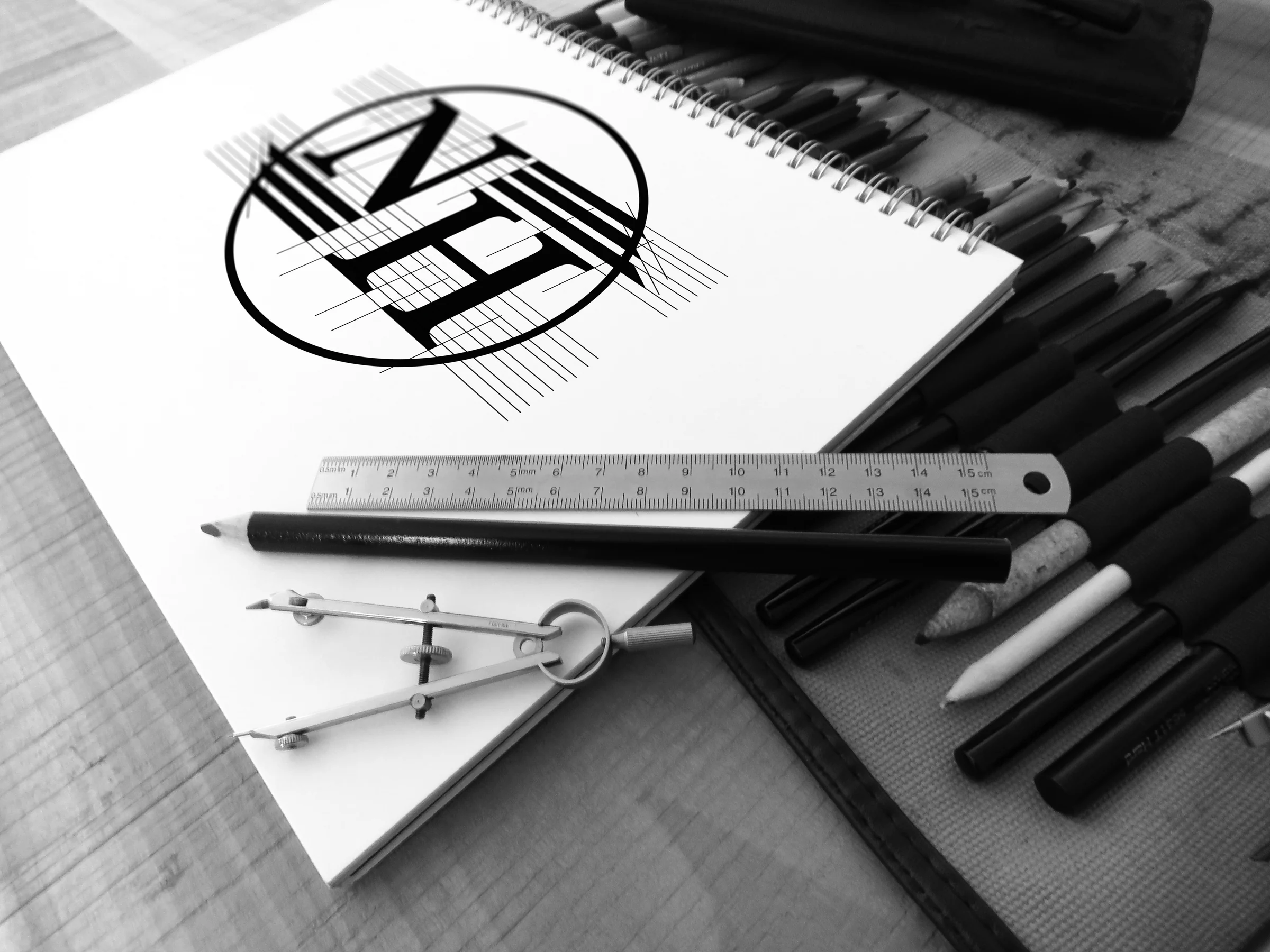Black-and-white photo of a spiral sketchbook with the NH monogram and its construction lines, a ruler, compass and pencils around it.