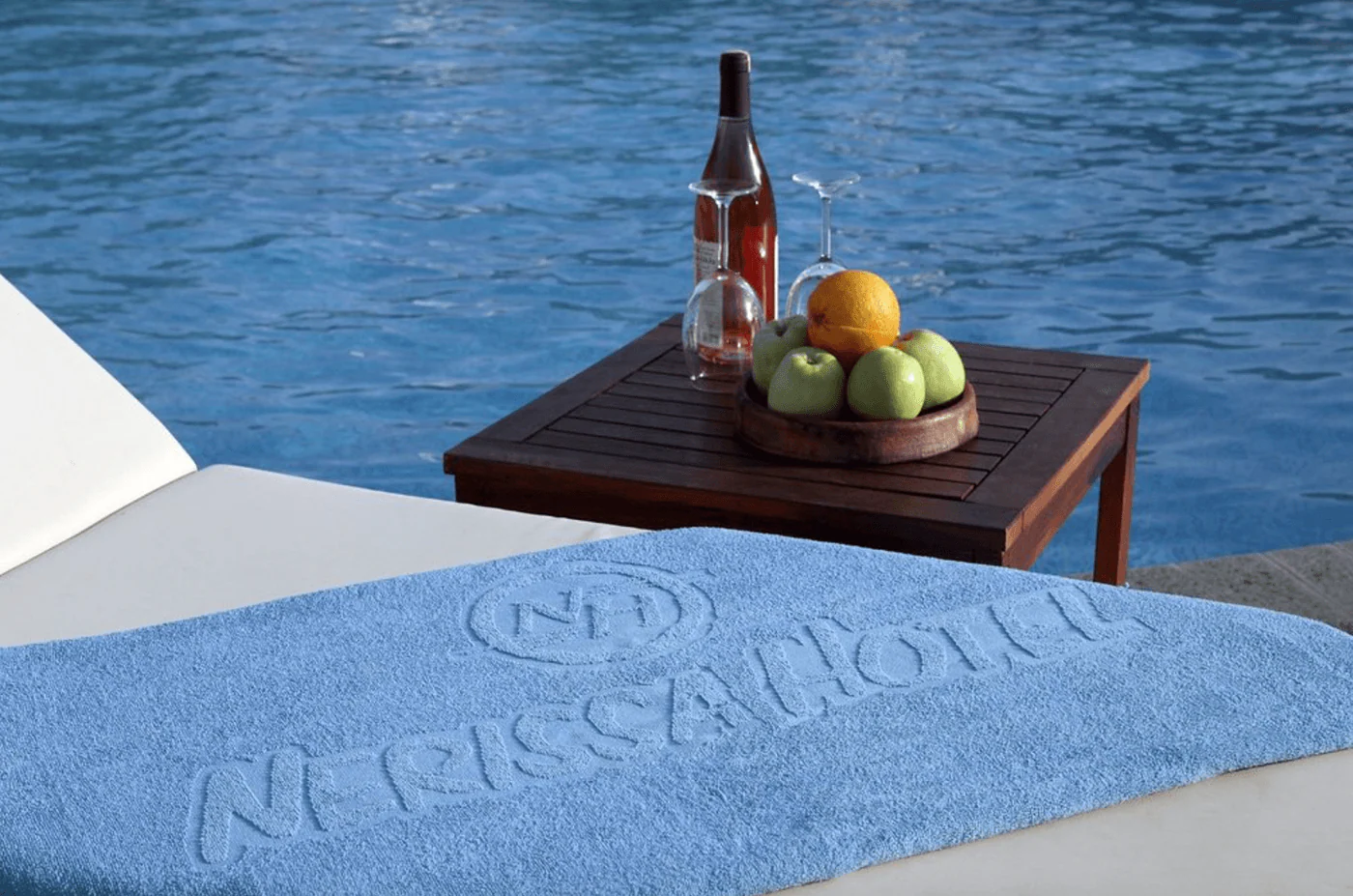 Light blue towel on a sunbed with a tone-on-tone NH mark and NERISSA HOTEL, by a poolside table with wine and fruit.