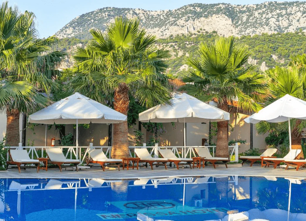 Pool lined with white parasols and loungers below a rocky, tree-covered mountain, the NH mark and hotel name on the pool floor.
