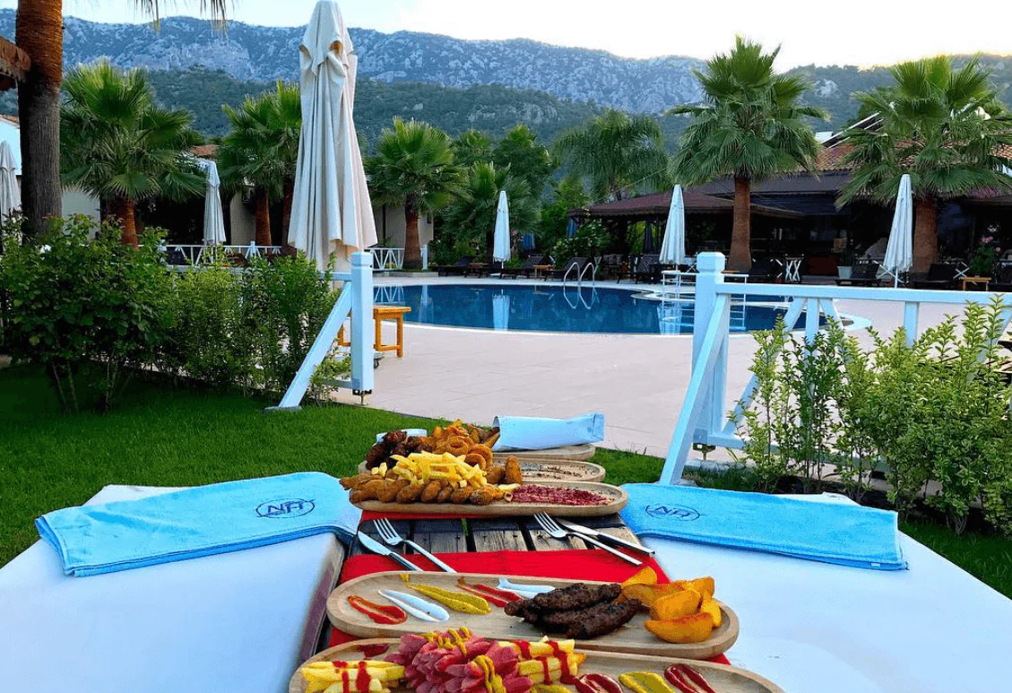 Food platters on a red runner between two sunbeds with folded light blue towels bearing the NH mark, a pool and palms behind.