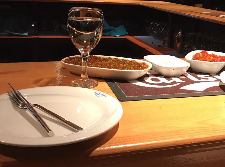 White plate with a small blue NH mark on the rim and cutlery on it, beside a stemmed glass and small dishes on a wooden bar.