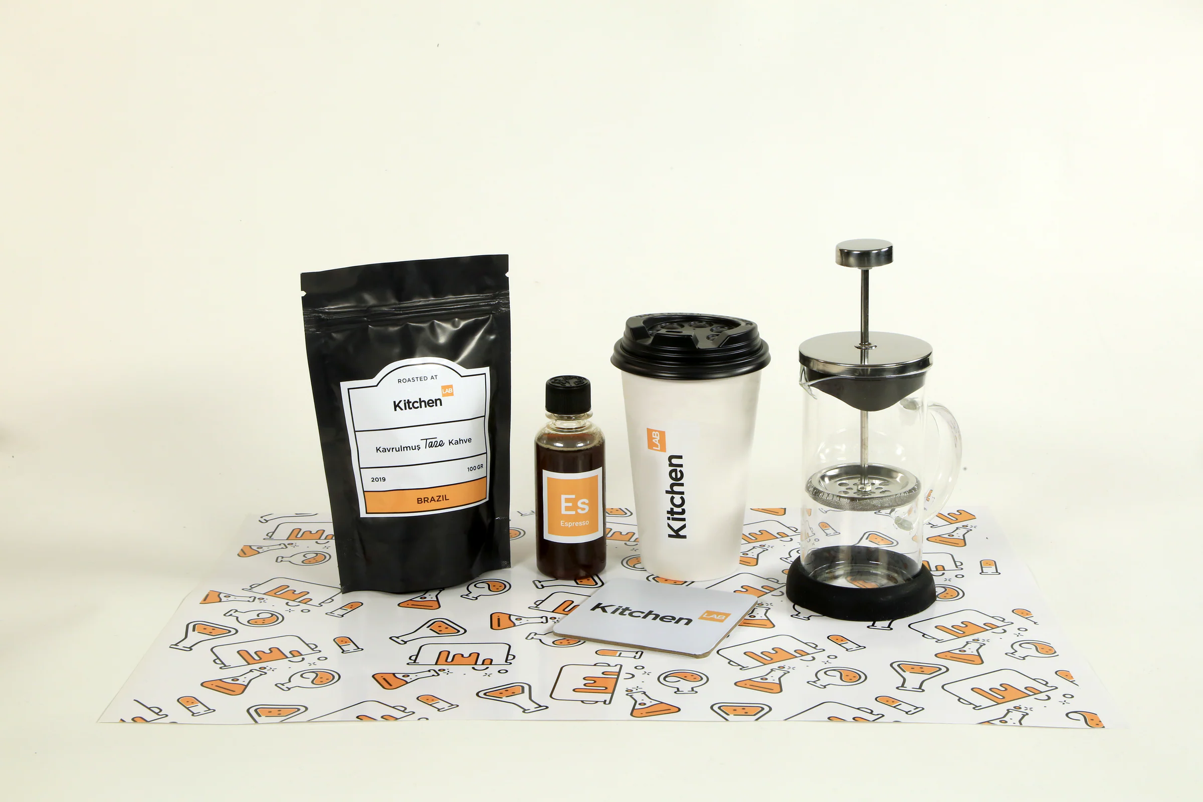 Studio shot of the KitchenLAB coffee pouch, espresso bottle, takeaway cup, coaster and French press on a patterned placemat.