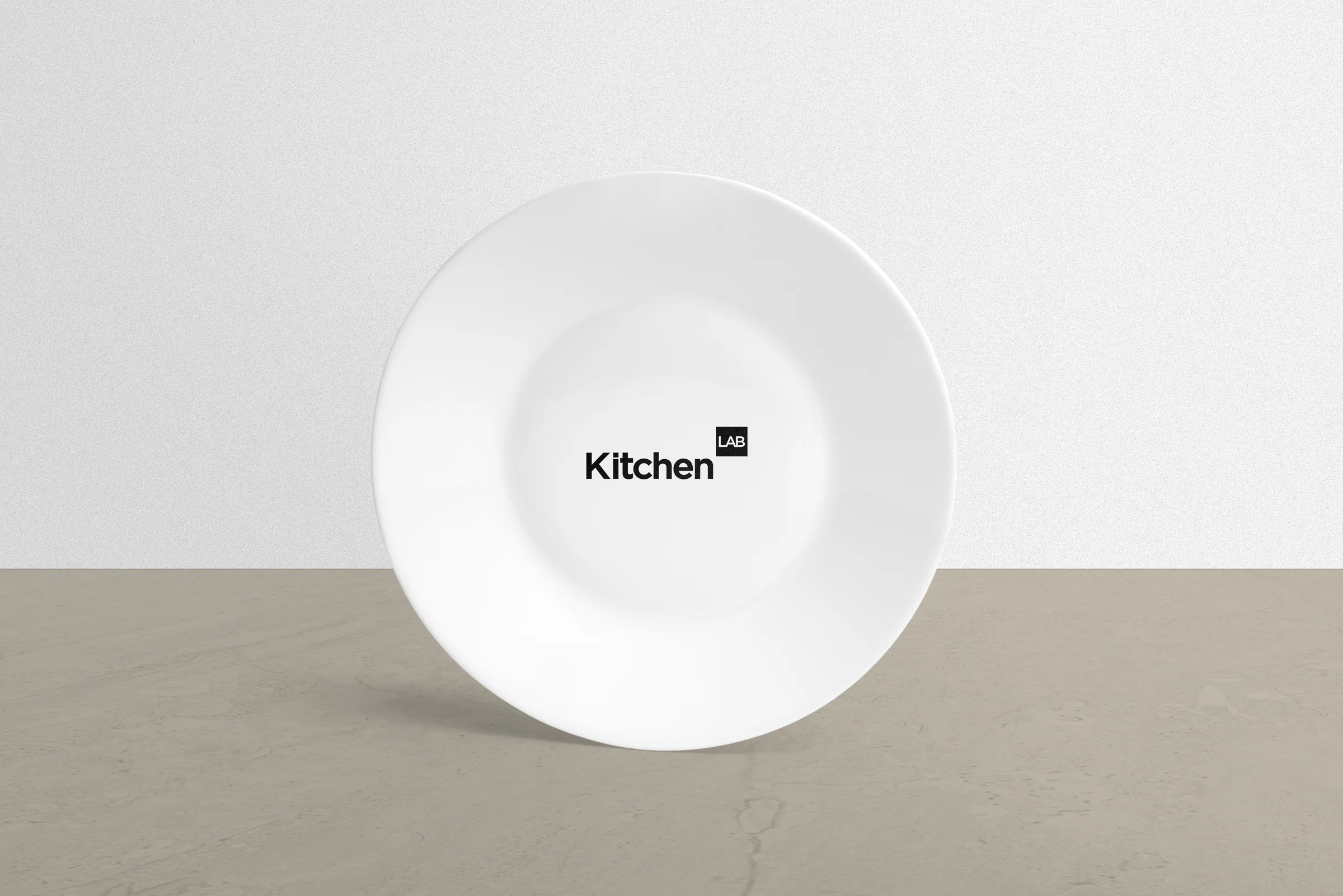 White plate with a small black KitchenLAB logo in the centre, leaning against a pale wall.