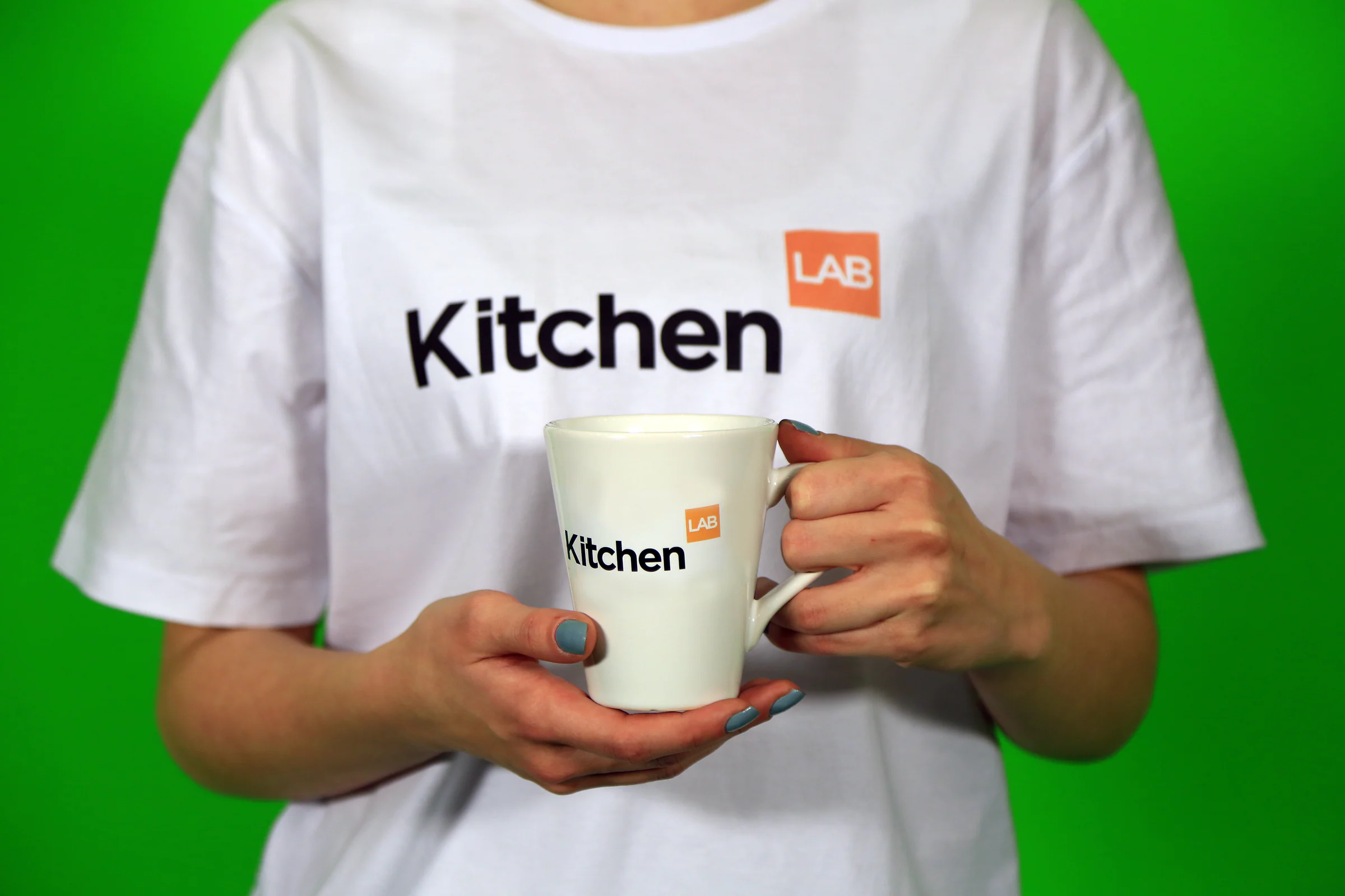 Person in a white KitchenLAB T-shirt holding a matching white logo mug against a green backdrop, face out of frame.