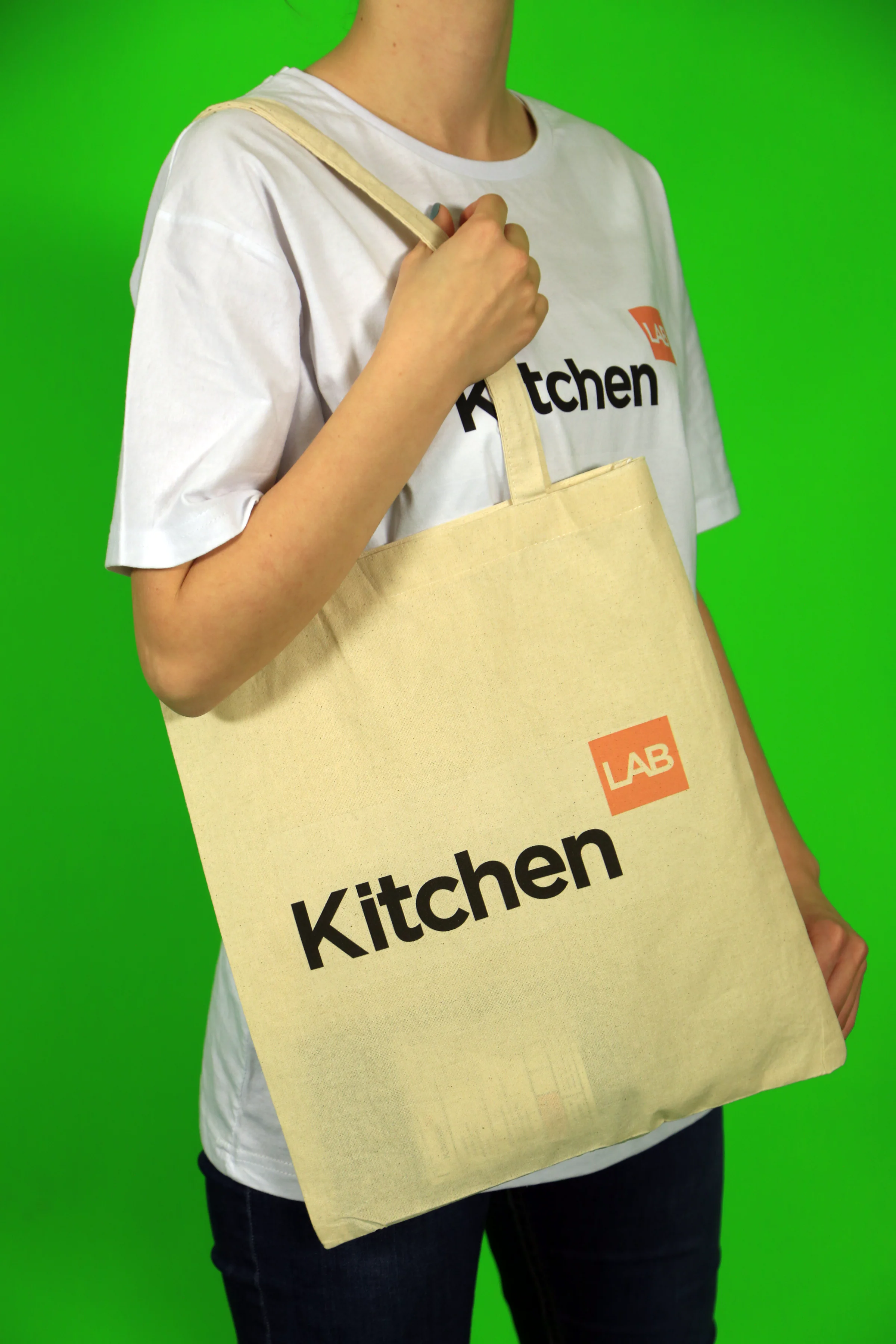 Person in a KitchenLAB T-shirt carrying a natural cotton tote bag printed with the logo, against a green backdrop.