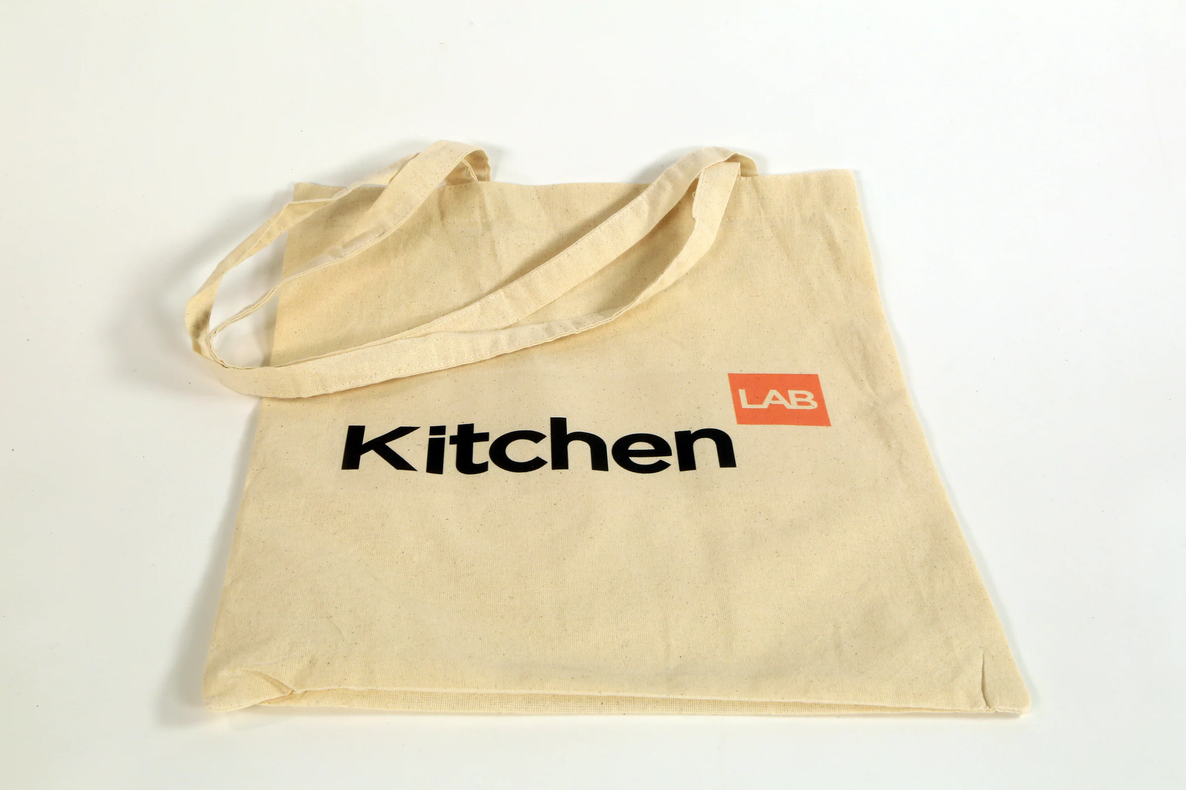 Natural cotton tote bag with the KitchenLAB logo in black and orange, laid flat on a white surface.