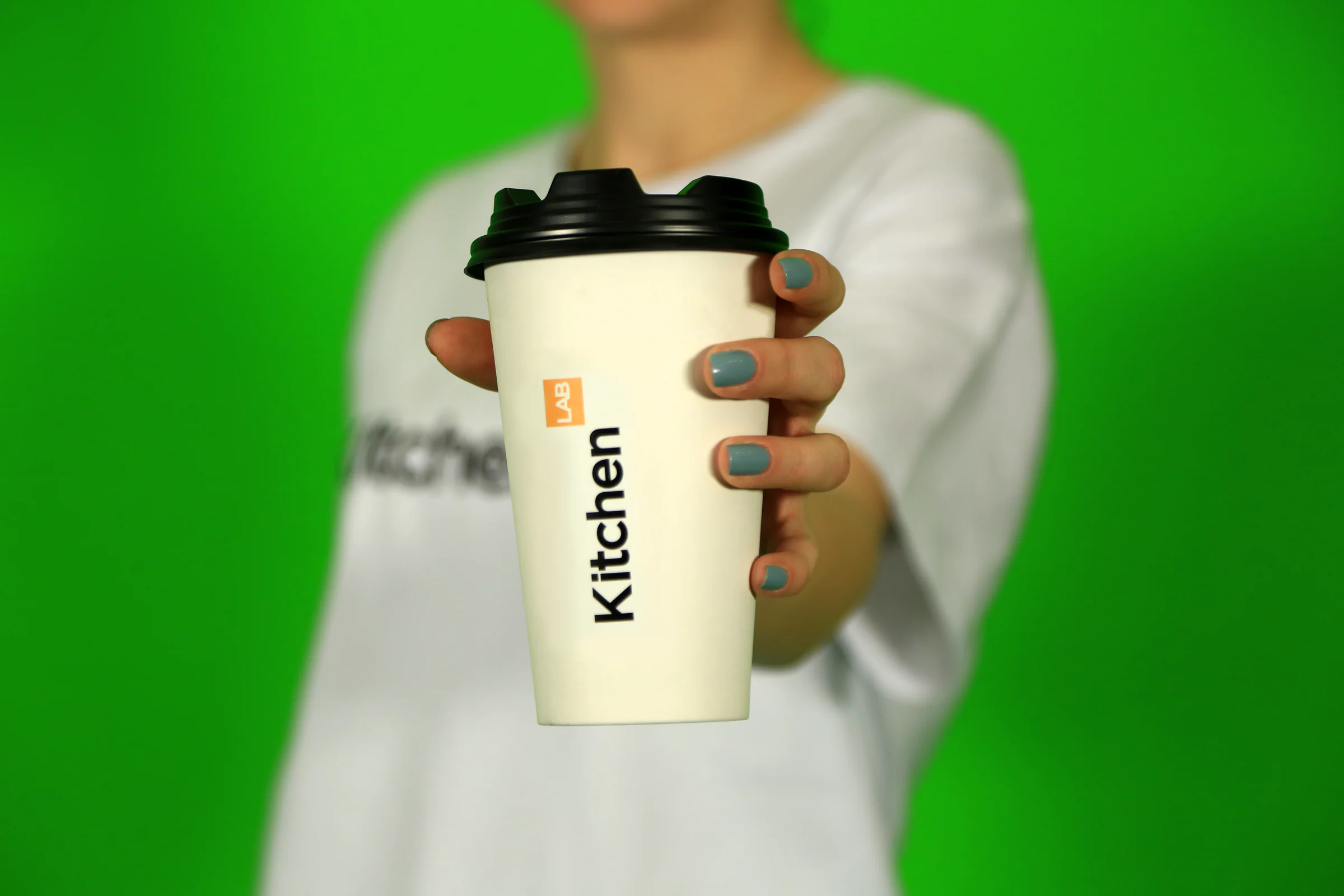 Hand holding out a white KitchenLAB takeaway cup with a black lid toward the camera, green backdrop behind.