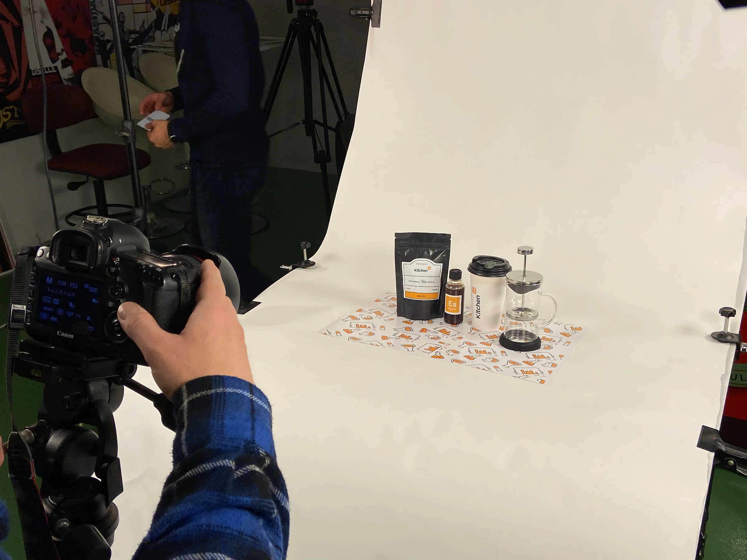 Backstage: a hand on a tripod-mounted camera frames the coffee set on a white sweep while another person stands behind.
