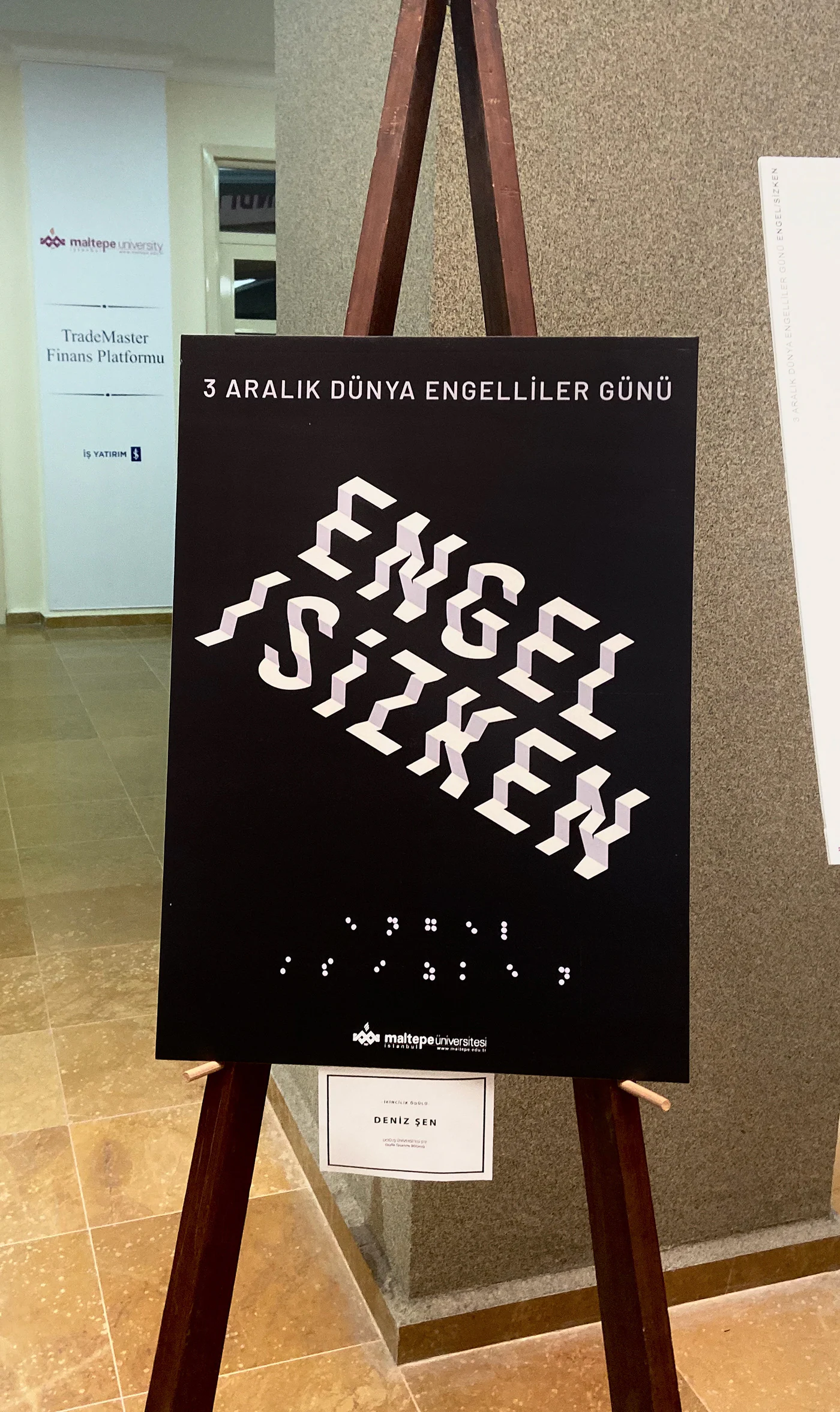 Printed poster headed 3 Aralık Dünya Engelliler Günü on a wooden easel in a hallway, with a Deniz Şen name card pinned below.